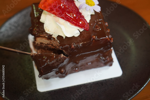 closeup chocolate cake decorated with a strawberry, a blueberry, and flowers and leaves, placed on a sheet of paper top a black ceramic plate, on brown wooden floor, brown wooden wall background