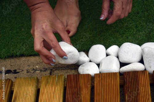 Close-up of a woman's hands arranging white decorative pebbles along a garden border beside a wooden deck and artificial grass. Outdoor landscaping and garden decoration concept.