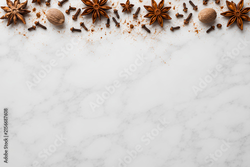Top view of aromatic winter spices including star anise, cloves, and nutmeg forming a top border on a white marble background with copy space.