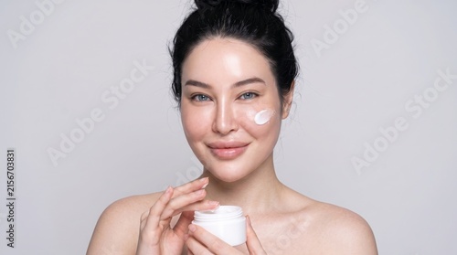 Woman applying skincare moisturizer with fresh radiant complexion against neutral background for beauty product advertisement
