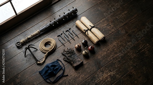 Ancient ninja tools including metal spikes iron club grappling hook paper scrolls leather gloves and dark mask on wooden floor