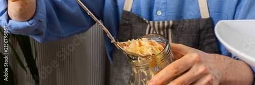 A 53-year-old man works in his kitchen fermenting cabbage and carrots. He carefully adds the mixture from a large bowl into a jar, showing concentration, banner