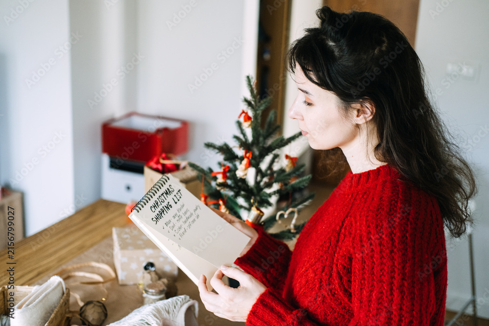 Fototapeta premium Woman turns page of notebook with CHRISTMAS SHOPPING LIST beside wrapped gifts and small tree. Personalized christmas gift list, gift ideas for each family member, thoughtful christmas gift planning
