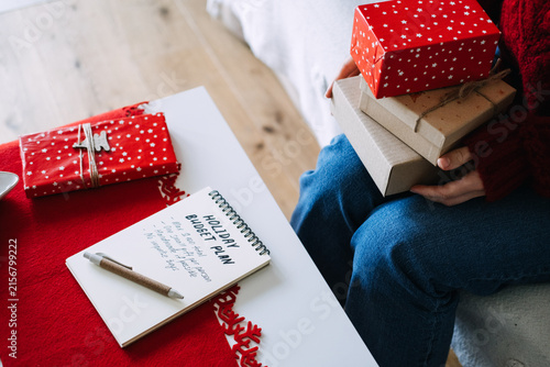 Woman holds stack of wrapped gifts beside notebook with HOLIDAY BUDGET PLAN on table. Christmas budget planning, mindful holiday spending, no impulse buying gift rules