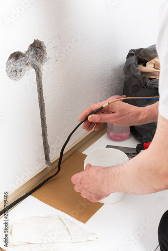 Worker runs new electrical wires into a cutout in a white apartment wall. This task prepares for the installation of an electrical outlet near the baseboard