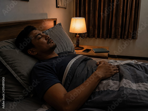 Man sleeping on his back under a blanket in a dark bedroom illuminated by a bedside lamp.generative AI