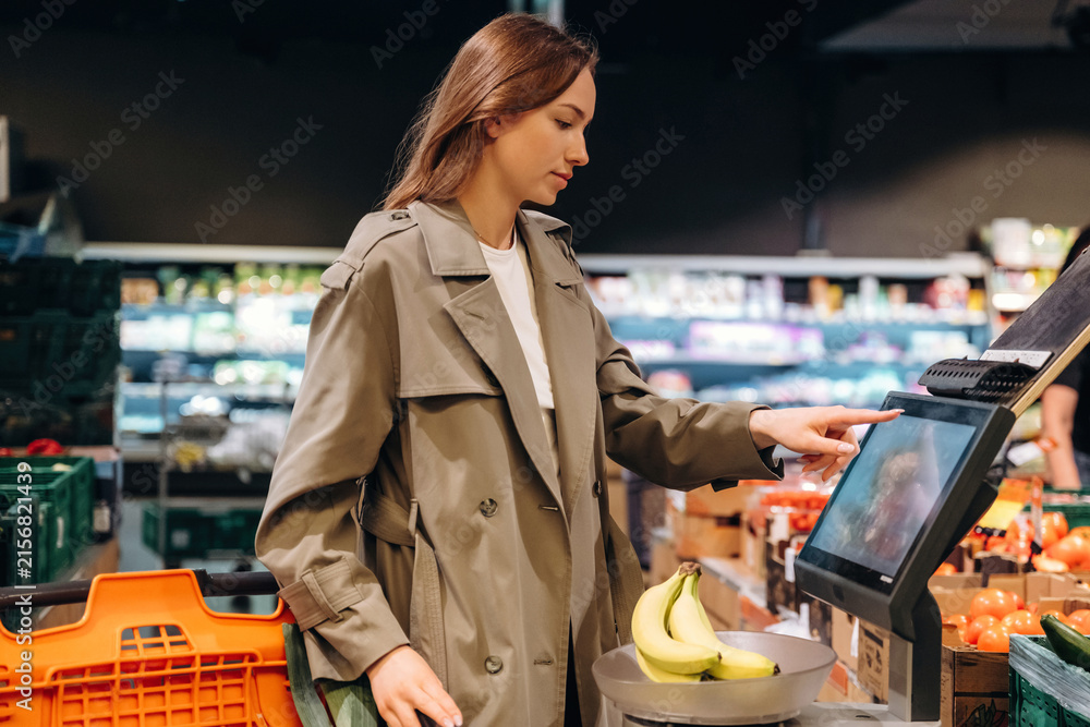 Naklejka premium Young woman in a beige trench coat using a self-service kiosk to check out groceries in a modern supermarket with fresh produce displayed in the background