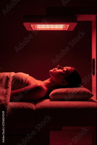 Woman lying down under red light therapy device for facial treatment. Use: spa brochure, beauty salon advertisement, wellness magazine.