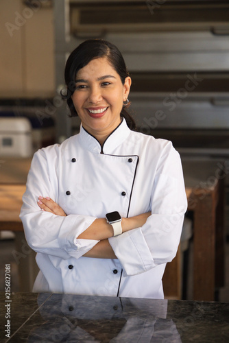 Mid-adult female chef standing behind stone counter in kitchen wearing chef coat and smartwatch