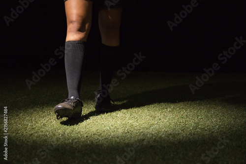 Female soccer player standing on turf at night wearing knee-high socks and cleats under spotlight