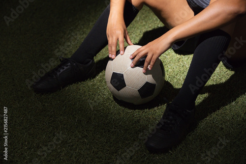 African American woman sitting on turf holding soccer ball at knees wearing socks shorts and cleats