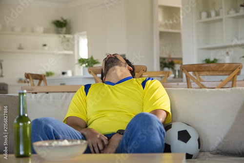 Photography Middle-aged man wearing yellow jersey slumping on sofa at home, soccer ball besi