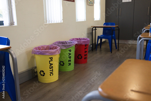 Three recycling bins are standing along classroom wall under windows, labeled PLASTIC PAPER GLASS