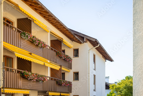 Photography A multi-story Alpine-style building featuring wooden balconies adorned with colorful flower boxes, yellow sun awnings, and overhanging wooden eaves against a clear sky
