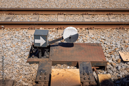 Canvas Print A manual railway switch indicator mechanism featuring a white directional arrow box and a black-and-white counterweight disc, positioned beside train tracks on gravel ballast