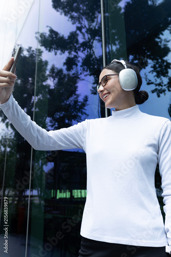 Vertical portrait of a confident Latina woman taking a selfie with her smartphone near a highly reflective glass building.