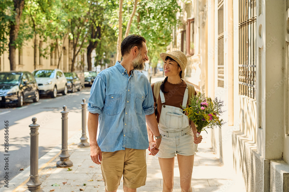Fototapeta premium Couple Walking Hand in Hand on Sunny Urban Street With Flowers and Smiles