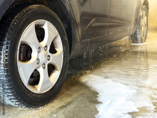 Close-up of a dark car wheel and side sill with water dripping on a wet concrete floor during a car wash.