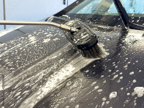 Washing a dark car hood with a soapy foam brush at a self-service car wash bay.