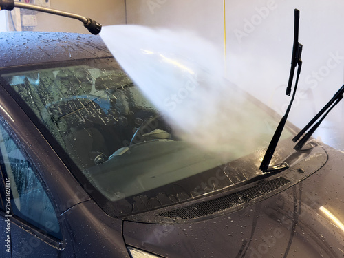 Power washing a dark car windshield with a high-pressure water sprayer at a self-service car wash.