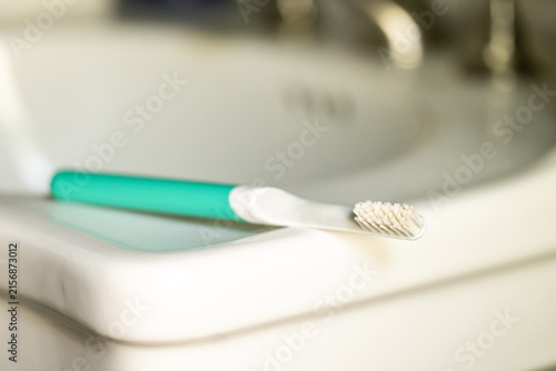 Bild auf Leinwand Green toothbrush rests on the edge of a white bathroom sink beside a chrome faucet, evoking daily oral care, personal routines, self-discipline, and healthy habits