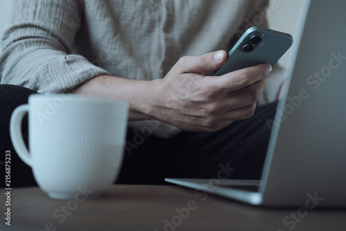 Close-up of a person using a smartphone while working on a laptop with coffee at a modern workspace.