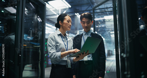 Asian Female Systems Analyst Using Laptop Computer to Review Diagnostic Results With Asian Male Infrastructure Engineer While Walking Through Server Rack Corridor in AI Data Center Facility.