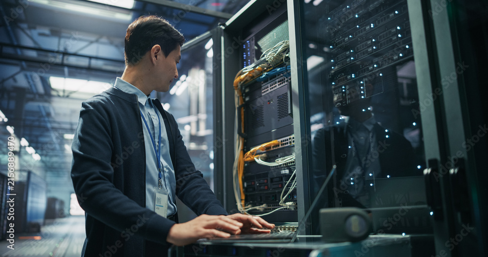 Fototapeta premium Asian Male It Support Technician With Id Badge Using Laptop Computer for Server Diagnostics and Network Troubleshooting Beside Open Rack Cabinet With Ethernet Cables in AI Data Center Facility