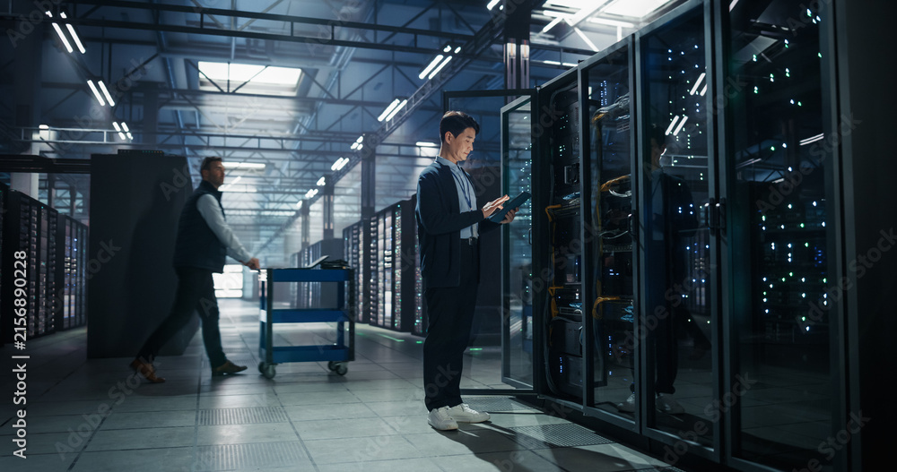 Fototapeta premium Asian Male Systems Administrator Holding Tablet Computer at Open Server Rack While Colleague Transports Hardware on Cart, Storage Capacity and Firmware Updates in AI Cloud Computing Data Center