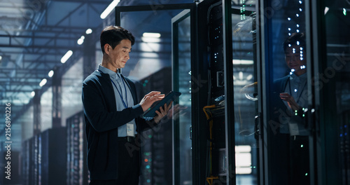 Asian Male It Specialist Using Tablet Computer to Configure Network Settings Beside Open Server Rack, Managing Data Migration and Uptime Analytics in AI Cloud Computing Colocation Facility