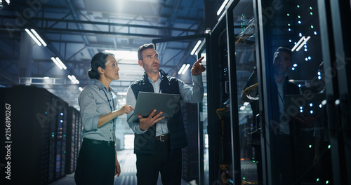 Caucasian Male Engineer Holding Laptop Computer Points up at Server Rack Hardware While Asian Female Supervisor Observes, Two Data Center Specialists Conducting Cloud Storage Capacity Audit