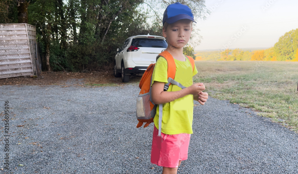 Obraz premium child standing on gravel path in calm rural scene with trees and field, side view, blue cap, yellow shirt, pink shorts, orange backpack, white car, wooden fence, serene mood.