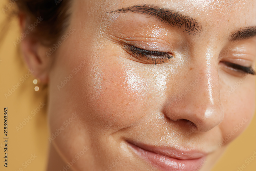 Obraz premium Glowing close up of oil sheen on cheekbone catching warm light, natural skin texture and freckles with soft smile and subtle eyeliner