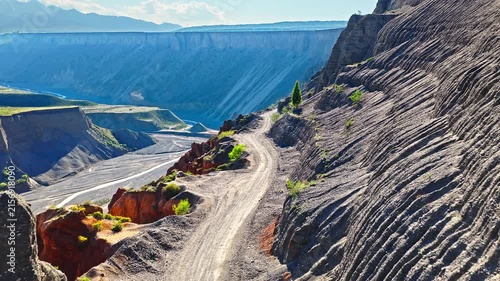 Aerial view of a winding dirt road along a steep eroded canyon cliff with layered geological strata formations in Anjihai Grand Canyon, Xinjiang, China