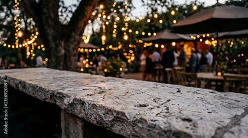 Close-up of textured stone ledge with blurred outdoor evening party ambiance, string lights, and people mingling, suitable for event promotion, hospitality marketing and social gathering imagery