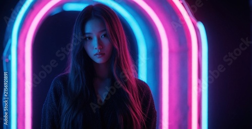 Young woman with long dark hair poses under colorful neon lights. Pink, blue glow illuminates face in dark studio. Cyberpunk style portrait features beautiful girl with moody expression. Fashion