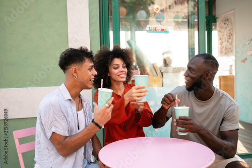 Diverse young friends enjoying contemporary cafe culture, laughing, toasting outside on a summer day