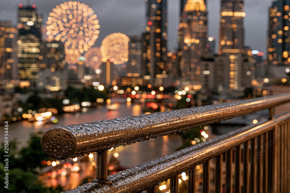 Fototapeta premium Raindrops on metal railing with city fireworks and night skyline view