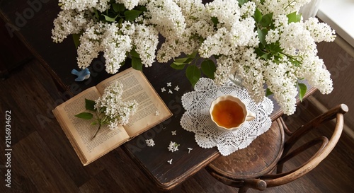 Rustic table with blooming white lilacs, an open book, and a cup of tea on a lace doily. Warm, nostalgic ambiance with natural light.
