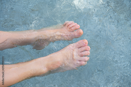 Top view of bare feet relaxing in clear, shallow turquoise ocean water. Soft focus on ripples and sunlight reflections, capturing a peaceful summer vacation lifestyle concept.