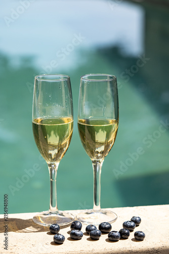 Two elegant glasses of white wine or champagne by a turquoise swimming pool with fresh blueberries. Refreshing summer lifestyle, soft focus, bright sunny mood and vacation vibes.