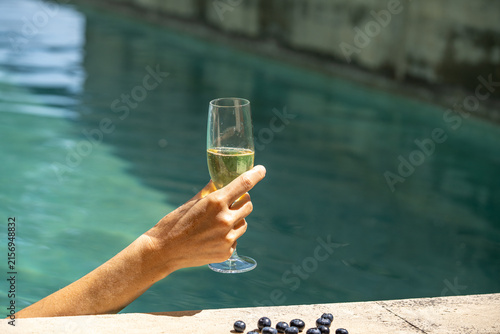 A hand holds a glass of sparkling white wine by a turquoise swimming pool. Sunny summer mood, refreshing lifestyle concept with soft focus and bright blue water texture.
