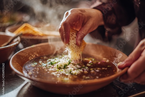 person hands dipping birria taco into rich soup with cheese melt, beef filling in hot consommé with steam in cooking environment, close up mexican cuisine preparation, restaurant
