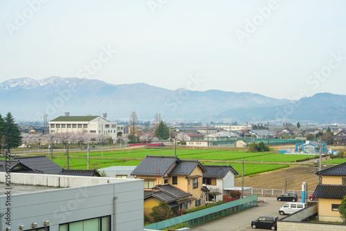 A high-angle landscape overview of a peaceful Japanese countryside town featuring residential houses, lush green agricultural fields, blooming sakura trees, and distant mountains under a soft hazy sky