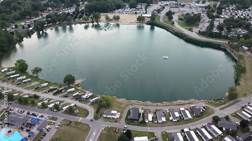 An aerial drone shot orbits above Sherkston Quarry, highlighting the floating inflatable water park obstacle course, the sandy beachfront, and the surrounding holiday RV campgrounds