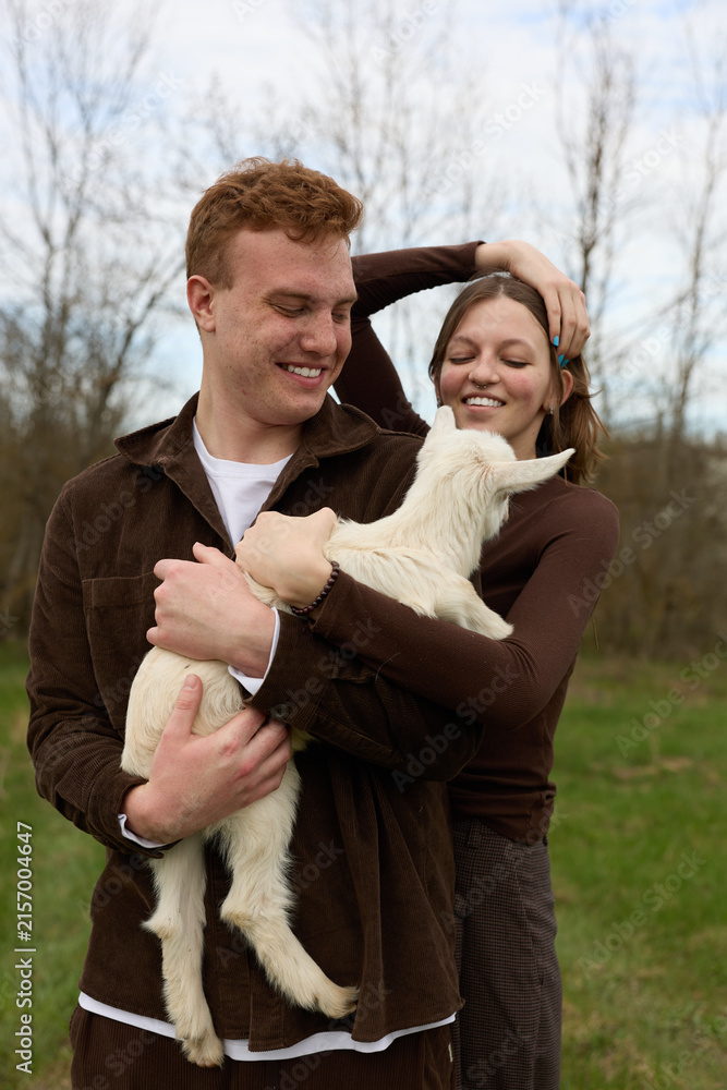 Naklejka premium Casual couple outdoors with a playful goat kid, warm smiles, and gentle touch, candid lifestyle moment, soft natural light, relaxed poses, rustic clothing, airy countryside background, wholesome