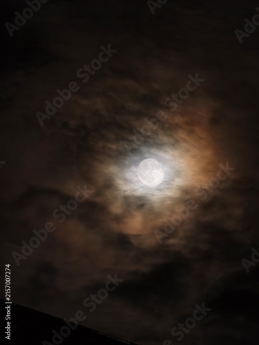 Bright full moon shining through dense, dark, and textured clouds in the night sky with a hint of orange
