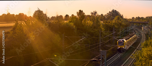 Canvas Print Intercity train traveling through a scenic railway corridor at sunset, surrounded by trees and overhead power lines