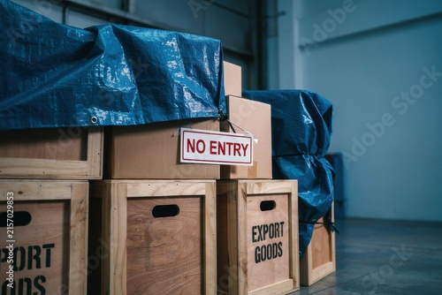 Stacked wooden crates and boxes covered with blue tarpaulins in a warehouse, with a red 'No Entry' sign.