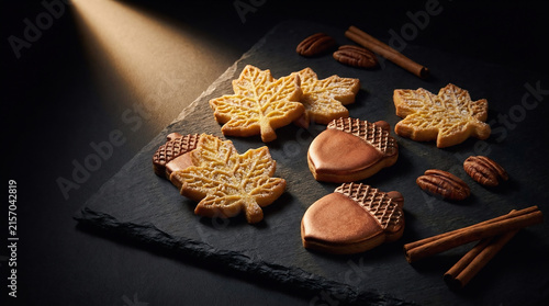 Autumn cookie designs featuring detailed leaf and acorn shapes on a textured black slate board with rustic cinnamon sticks and pecan nuts on the table
